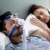 sleep apnea heart health risks shown by man sleeping with CPAP mask beside partner in bed