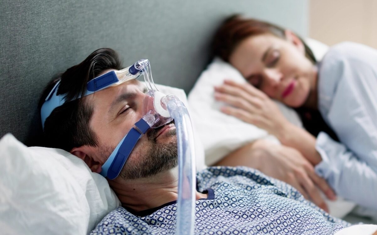 sleep apnea heart health risks shown by man sleeping with CPAP mask beside partner in bed