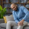 Man sitting on couch grimacing in knee pain considering when to see a doctor for pain management evaluation