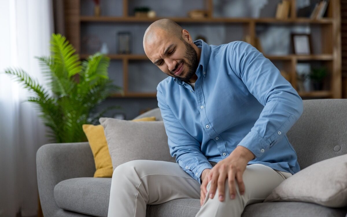 Man sitting on couch grimacing in knee pain considering when to see a doctor for pain management evaluation